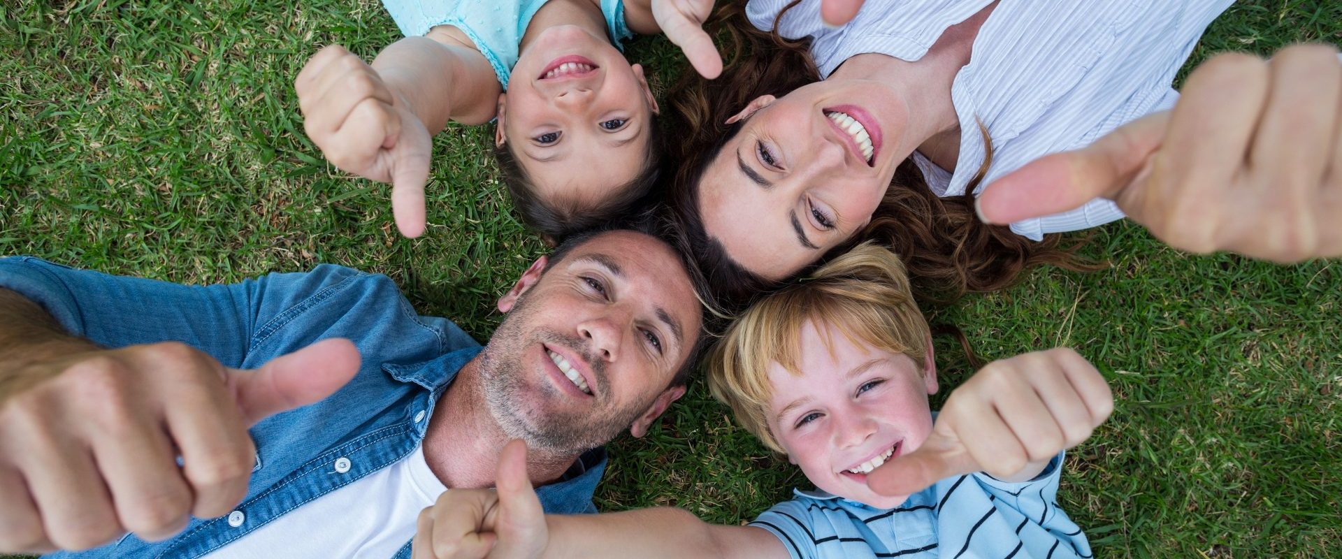 Familie mit Mutter, Vater, Tochter und Sohn liegen im Gras und schauen nach oben. Sie halten ihre Hände in die Luft. Sie strecken ihre Daumen nach oben.