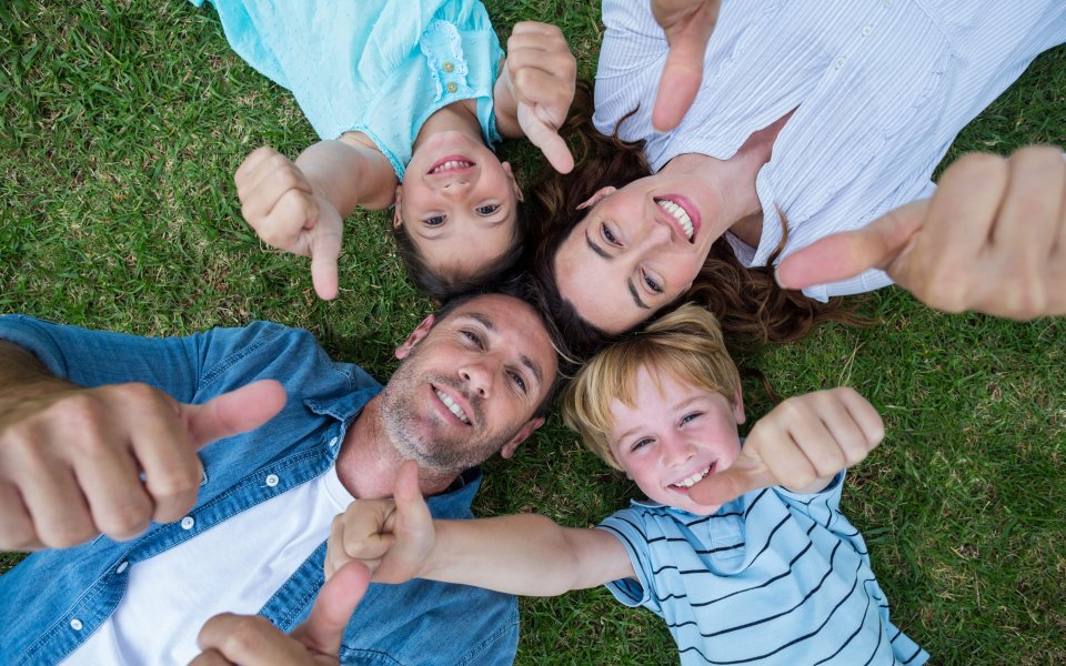 Familie mit Mutter, Vater, Tochter und Sohn liegen im Gras und schauen nach oben. Sie halten ihre Hände in die Luft. Sie strecken ihre Daumen nach oben.