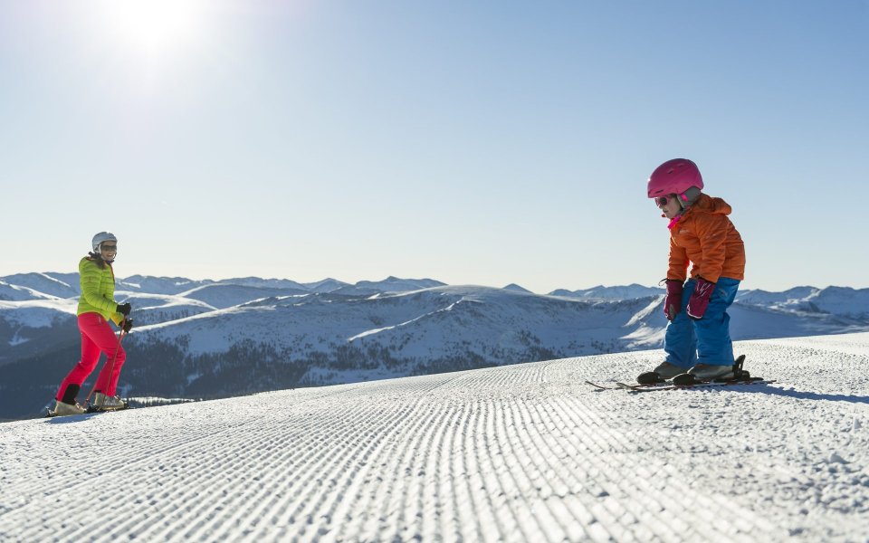 kleines Kind fährt mit gebeugten Oberkörper und den Händen auf den Knien auf der Piste Schi. Eine Frau steht unten weiter auf der Piste.