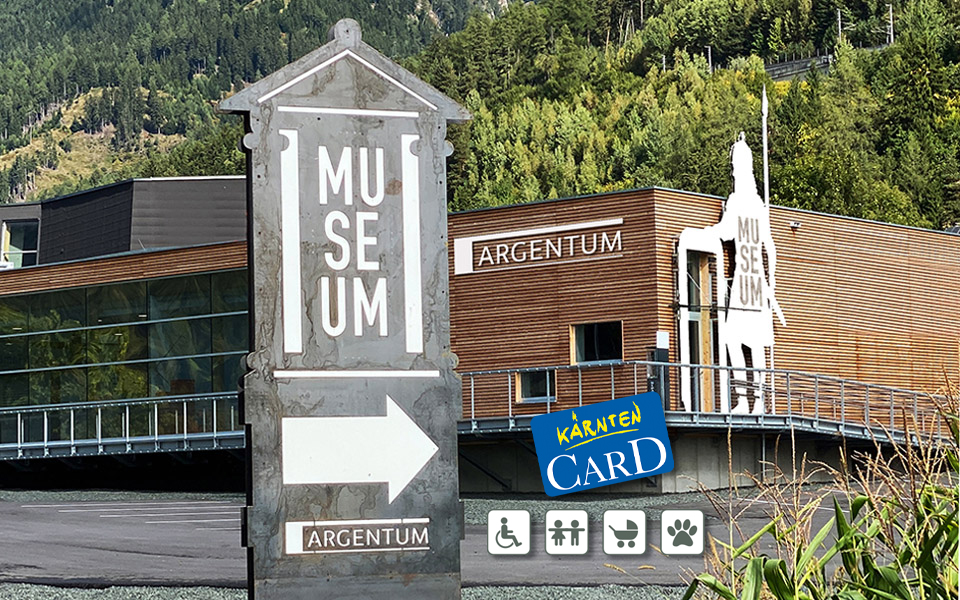 Zu sehen ist die Vorderansicht des Museum mit dem sechs Meter großen Keltenkrieger, der rechts von der Eingangstür angebracht ist. Zudem sieht man im Vordergrund eine Museumsankündigungstafel sowie Symbole für die Kärnten Card, Barrierefreiheit, familienfreundlich, Hunde erlaubt und Kinderwagen-gerecht.